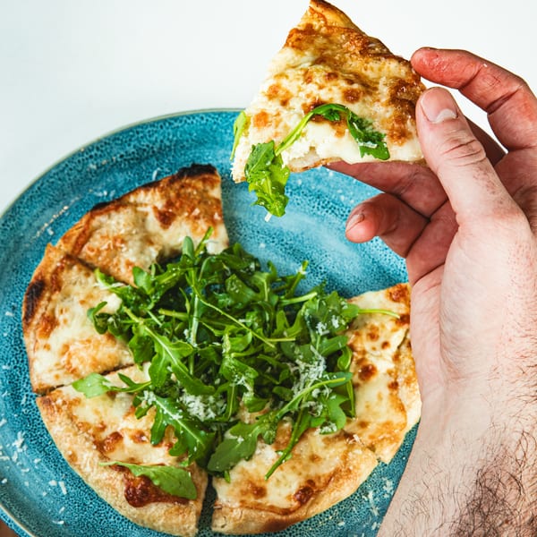 A hand holding a slice of flatbread pizza with melted cheese and arugula over a blue plate.